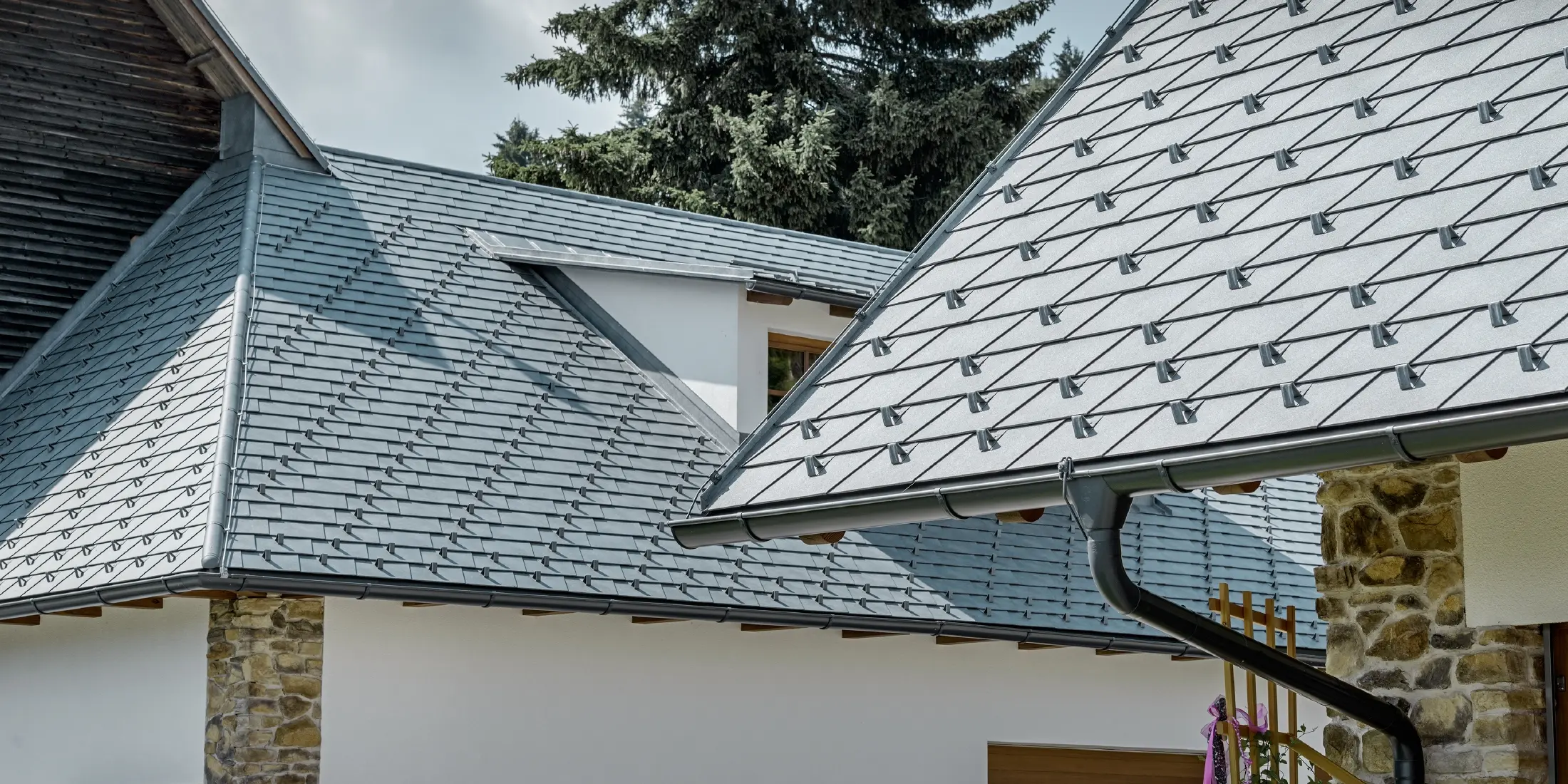 Detailaufnahme einer Alu-Dacheindeckung von PREFA; Dachschindel in Steingrau mit der Alu-Dachrinne in Anthrazit von PREFA; Im Hintergrund erkennt man eine Schleppgaube mit Stehfalzdach. Die Fassade ist weiß mit eingearbeiteten Steinelementen.