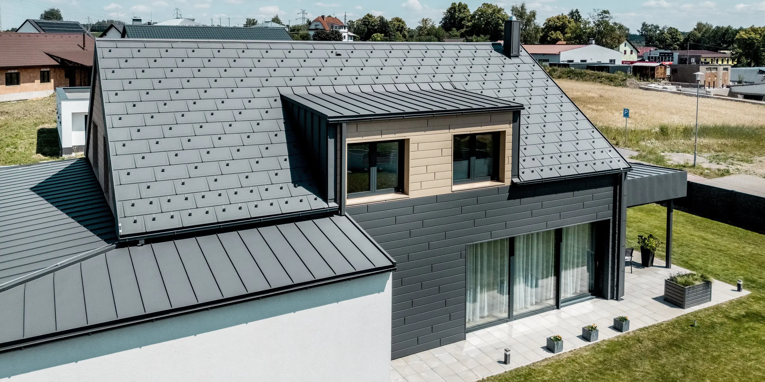 Moderne Ansicht des Einfamilienhauses mit PREFA Aluminiumlösungen an Dach und Fassade. Die PREFA Dachplatte R.16 und PREFALZ in P.10 Dunkelgrau verbinden sich mit den Sidings in Dunkelgrau und Sandbraun zu einer klaren, harmonischen Gebäudehülle. Großzügige Fensterflächen und die offene Terrasse unterstreichen die zeitgemäße Architektur, während die langlebigen Aluminiumprodukte zuverlässigen Schutz und dauerhafte Schönheit bieten.