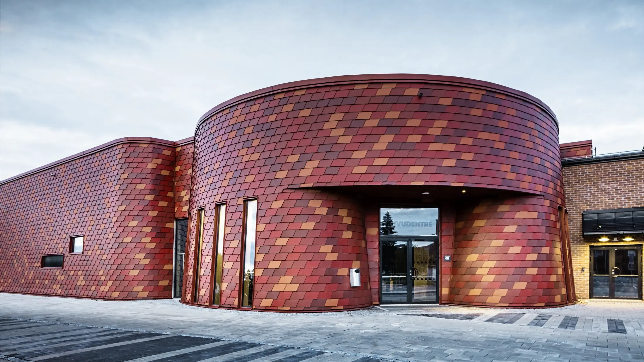 Eishalle mit Flachdach und geschwungener Fassade, die Fassade ist mit Wandschindeln aus Aluminium von PREFA in verschiedenen Rottönen verkleidet - Oxydrot, Ziegelrot, Rotbraun, Braunrot