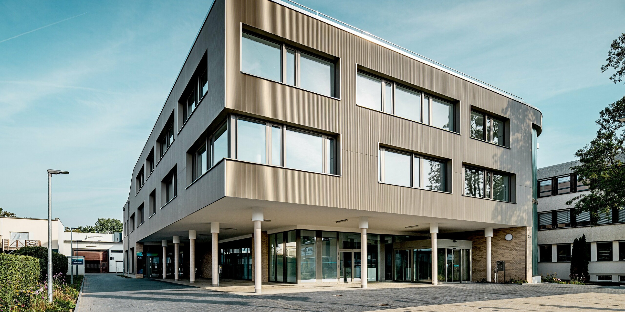 Das Bild zeigt das moderne Festo-Gebäude in Langenhagen, dessen beeindruckende Fassade mit PREFA Produkten in Bronze optisch hervortritt. Die klare Struktur der Strangpressprofile in Zackenform verleiht der Fassade eine dynamische Textur, die in Kombination mit den eleganten Aluminium Verbundplatten auf der gegenüberliegenden Seite zu einem stimmigen Gesamtkonzept führt. Besonders auffällig ist die markante Rundung der Gebäudeecke, die dem Bauwerk eine elegante Leichtigkeit verleiht. Dank der PREFA Materialien ist die Fassade nicht nur ein Blickfang, sondern auch langlebig, pflegeleicht und beständig gegenüber Witterungseinflüssen – ideale Voraussetzungen für ein zukunftsfähiges Bauwerk.