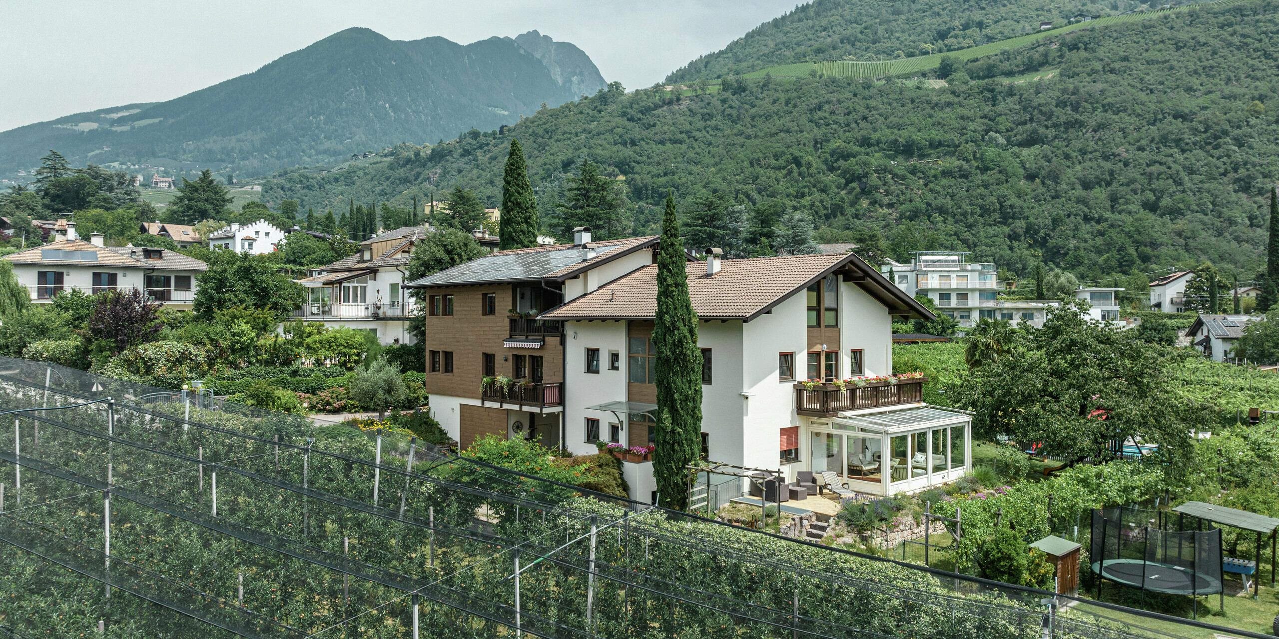 Luftaufnahme der Ferienwohnungen Prechtlhof in Meran, Südtirol, umgeben von Obstgärten und einer malerischen Berglandschaft. Die Fassade wurde mit PREFA Sidings in Walnussbraun modernisiert, wodurch eine langlebige und wartungsfreie Holzoptik entsteht, die sich harmonisch in die alpine Umgebung einfügt.