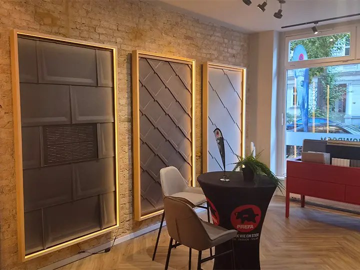 Innenansicht des PREFA Showrooms in Berlin mit beleuchteten Produktwänden mit verschiedenen PREFA-Fassadensystemen sowie einzelnen Produktmustern und einem Beratungsbereich mit Sitzgelegenheiten.