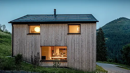 Das minimalistische Holzhaus Immi in Ebnit leuchtet am bewaldeten Berghang in der Abenddämmerung. Auf dem klar geformten Satteldach verschmelzen schwarze PREFA Solardachplatten nahezu unsichtbar mit der Dachplatte R.16 in P.10 Schwarz. Die robuste, witterungsbeständige Aluminium-Dachhaut erzeugt Solarstrom, schützt das Gebäude dauerhaft und unterstreicht die ruhige, moderne Architektur des alpinen Holzhauses.