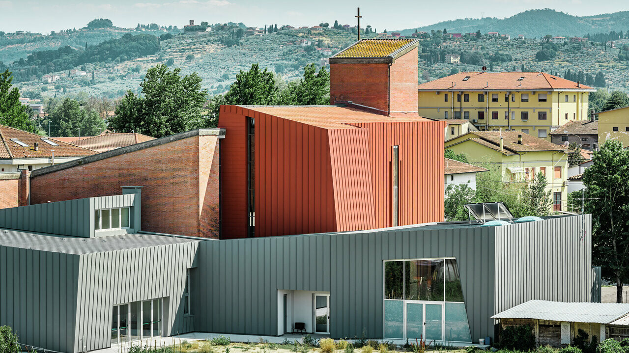 Die Kirche in Quarrata, Italien, ist mit PREFALZ in den Farben P.10 Hellgrau und P.10 Ziegelrot eingedeckt. Das Gebäude vereint moderne und traditionelle Elemente, wobei der obere Teil in Ziegelrot und der untere Teil in Hellgrau gestaltet ist. Die Kirche befindet sich in einer malerischen Umgebung mit Hügeln und grüner Landschaft im Hintergrund. In der Nähe stehen weitere Gebäude in verschiedenen Farben, und der Himmel ist klar mit wenigen Wolken.